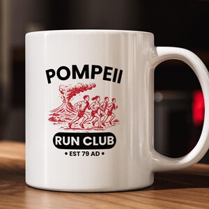 May include: White ceramic mug with the words "Pompeii Run Club" in black, and a red graphic of runners and a volcano. The mug also includes the text "Est 79 AD".