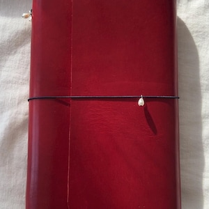Op de afbeelding: Een rood leren notitieboek met een omslag en een donkere koordsluiting. Het notitieboek is gesloten, met een kleine, decoratieve kraal aan het koord. Het leer heeft een gladde, licht glanzende afwerking. Het notitieboek is rechthoekig.