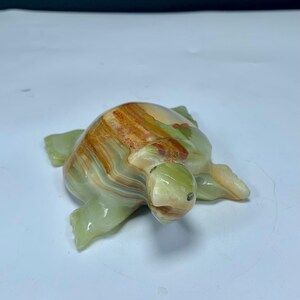 May include: A polished stone turtle figurine featuring a smooth, translucent surface with banded layers of light green, cream, and deep amber-brown colors. The sculpture displays a rounded shell and four flippers, with a small head extending forward.