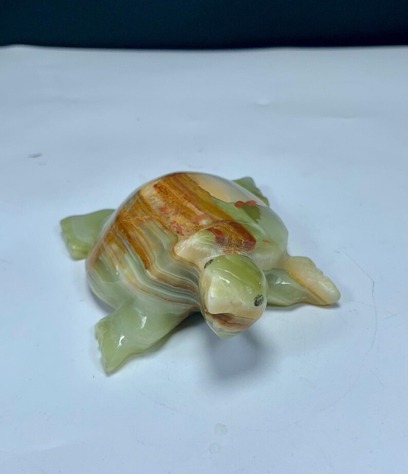 May include: A polished stone turtle figurine featuring a smooth, translucent surface with banded layers of light green, cream, and deep amber-brown colors. The sculpture displays a rounded shell and four flippers, with a small head extending forward.
