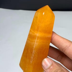 May include: A polished orange crystal tower with a hexagonal shape and a pointed top. The stone features translucent layers and natural internal banding throughout its structure, displaying a vibrant, warm, and saturated golden-orange color.
