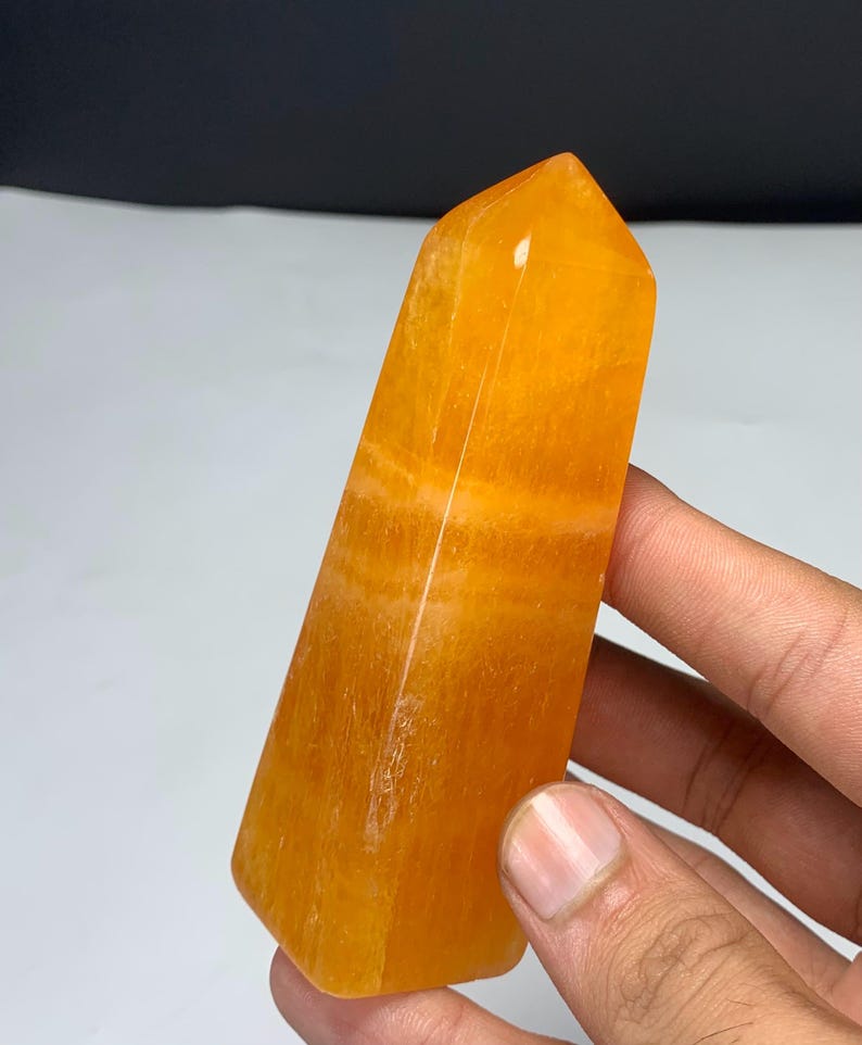 May include: A polished orange crystal tower with a hexagonal shape and a pointed top. The stone features translucent layers and natural internal banding throughout its structure, displaying a vibrant, warm, and saturated golden-orange color.