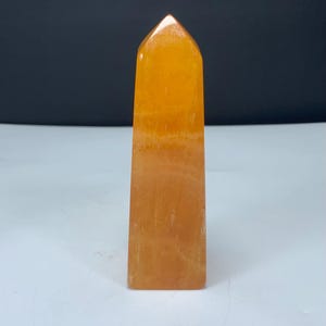 May include: A polished orange crystal tower with a pointed hexagonal top and a smooth, translucent surface. The stone features natural color variations ranging from deep amber to lighter golden hues, standing upright with a flat base.