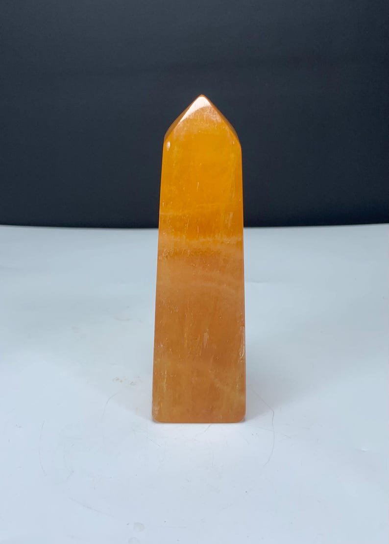 May include: A polished orange crystal tower with a pointed hexagonal top and a smooth, translucent surface. The stone features natural color variations ranging from deep amber to lighter golden hues, standing upright with a flat base.