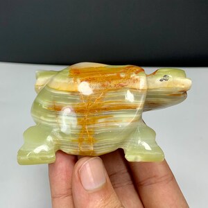 May include: A polished stone turtle figurine carved from banded green and orange onyx. The sculpture features a smooth, glossy surface with natural horizontal layers of translucent light green and vibrant orange mineral deposits throughout the body.