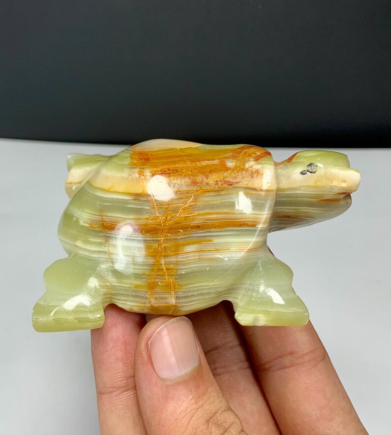 May include: A polished stone turtle figurine carved from banded green and orange onyx. The sculpture features a smooth, glossy surface with natural horizontal layers of translucent light green and vibrant orange mineral deposits throughout the body.
