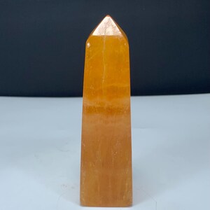 May include: A polished orange crystal tower with a pointed top and a rectangular base. The stone features a translucent quality with visible internal mineral inclusions and natural variations in the vibrant orange color throughout the structure.