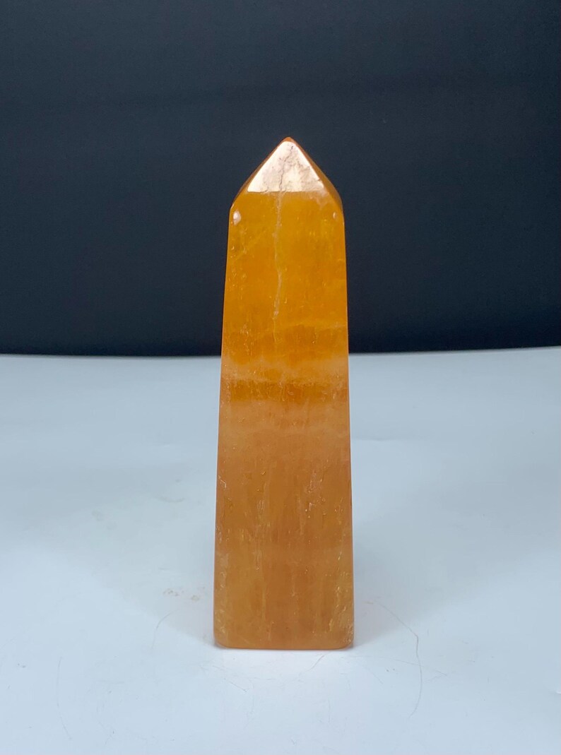 May include: A polished orange crystal tower with a pointed top and a rectangular base. The stone features a translucent quality with visible internal mineral inclusions and natural variations in the vibrant orange color throughout the structure.