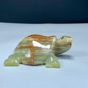 May include: A polished stone turtle figurine carved from banded green and brown onyx. The sculpture features horizontal layers of translucent light green and earthy brown mineral veins, with a smooth, glossy finish and a small dark eye detail.
