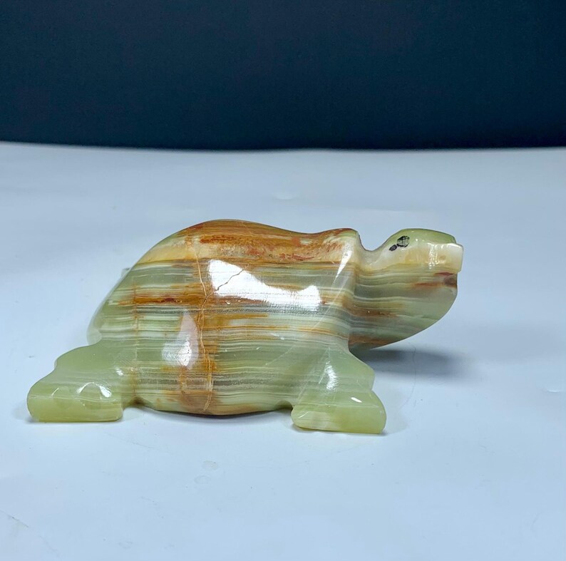 May include: A polished stone turtle figurine carved from banded green and brown onyx. The sculpture features horizontal layers of translucent light green and earthy brown mineral veins, with a smooth, glossy finish and a small dark eye detail.