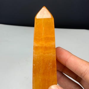 May include: A polished orange crystal tower with a pointed top and a rectangular base. The stone features a translucent, vibrant orange hue with visible internal mineral inclusions and natural banding patterns throughout the vertical structure.