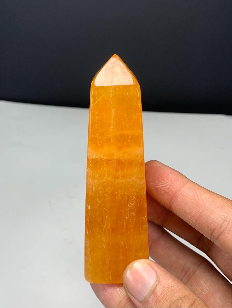May include: A polished orange crystal tower with a pointed top and a rectangular base. The stone features a translucent, vibrant orange hue with visible internal mineral inclusions and natural banding patterns throughout the vertical structure.