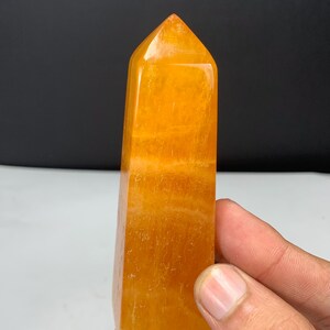 May include: A polished orange calcite crystal tower with a pointed top and a rectangular base. The stone features a vibrant, translucent orange hue with visible internal banding and natural mineral inclusions throughout its smooth surface.