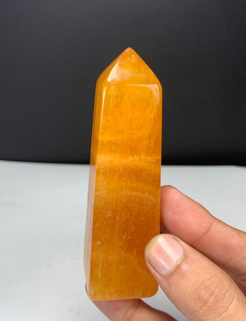 May include: A polished orange calcite crystal tower with a pointed top and a rectangular base. The stone features a vibrant, translucent orange hue with visible internal banding and natural mineral inclusions throughout its smooth surface.