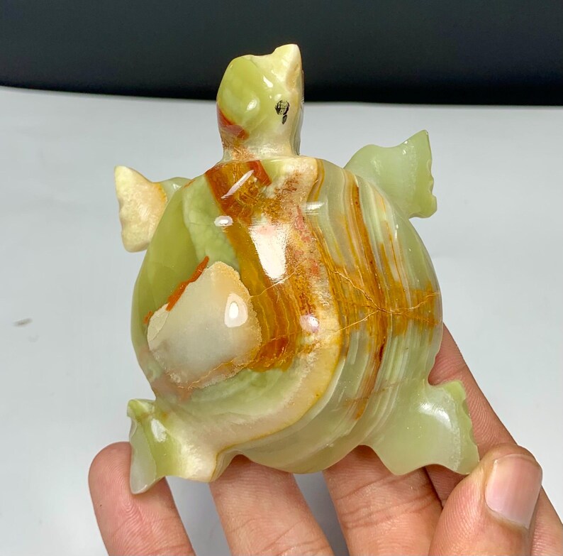 May include: A polished turtle figurine carved from light green and translucent banded stone. The sculpture features a smooth, glossy finish with prominent orange and brown mineral veins running across the shell and head, showcasing natural stone patterns.