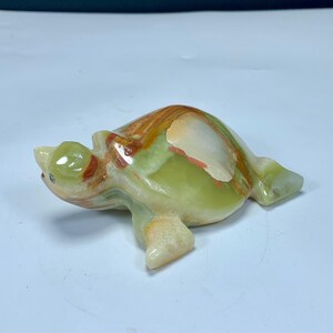 May include: A polished stone turtle figurine carved from translucent green onyx with natural banding in shades of cream, tan, and rust orange. The smooth, rounded shell features distinct mineral layers, while the head and flippers are sculpted.