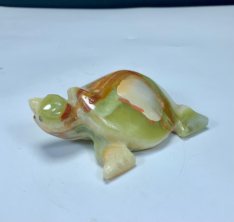 May include: A polished stone turtle figurine carved from translucent green onyx with natural banding in shades of cream, tan, and rust orange. The smooth, rounded shell features distinct mineral layers, while the head and flippers are sculpted.