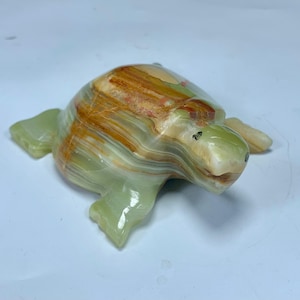 May include: A carved stone turtle figurine featuring a polished surface with banded layers of translucent light green, cream, and deep amber brown colors. The sculpture displays a smooth, rounded shell and defined flippers, showcasing the natural mineral patterns and variations inherent in the stone material.