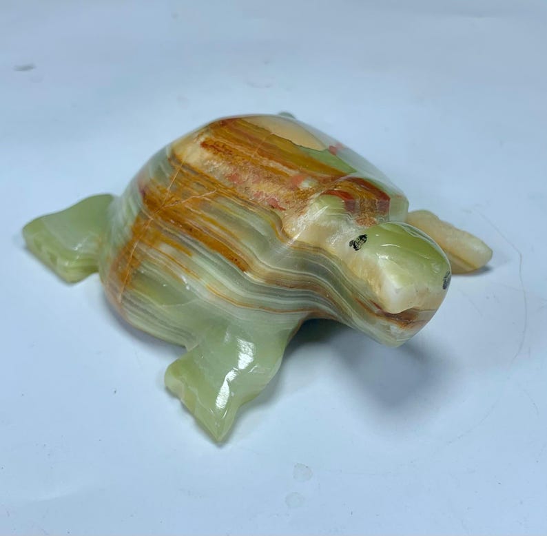 May include: A carved stone turtle figurine featuring a polished surface with banded layers of translucent light green, cream, and deep amber brown colors. The sculpture displays a smooth, rounded shell and defined flippers, showcasing the natural mineral patterns and variations inherent in the stone material.