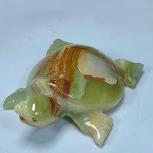 May include: A polished stone turtle figurine carved from banded onyx. The sculpture features a smooth, glossy surface with translucent light green, cream, and deep orange-brown mineral layers that create a natural, swirling pattern across the shell.