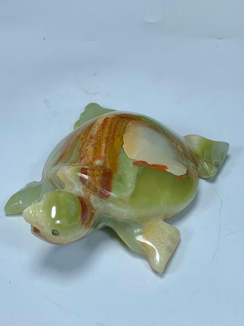 May include: A polished stone turtle figurine carved from banded onyx. The sculpture features a smooth, glossy surface with translucent light green, cream, and deep orange-brown mineral layers that create a natural, swirling pattern across the shell.