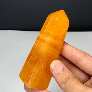 May include: A polished orange crystal tower with a hexagonal base and a pointed top. The stone features a translucent, vibrant orange hue with visible internal banding and natural mineral inclusions throughout its smooth, faceted surface.