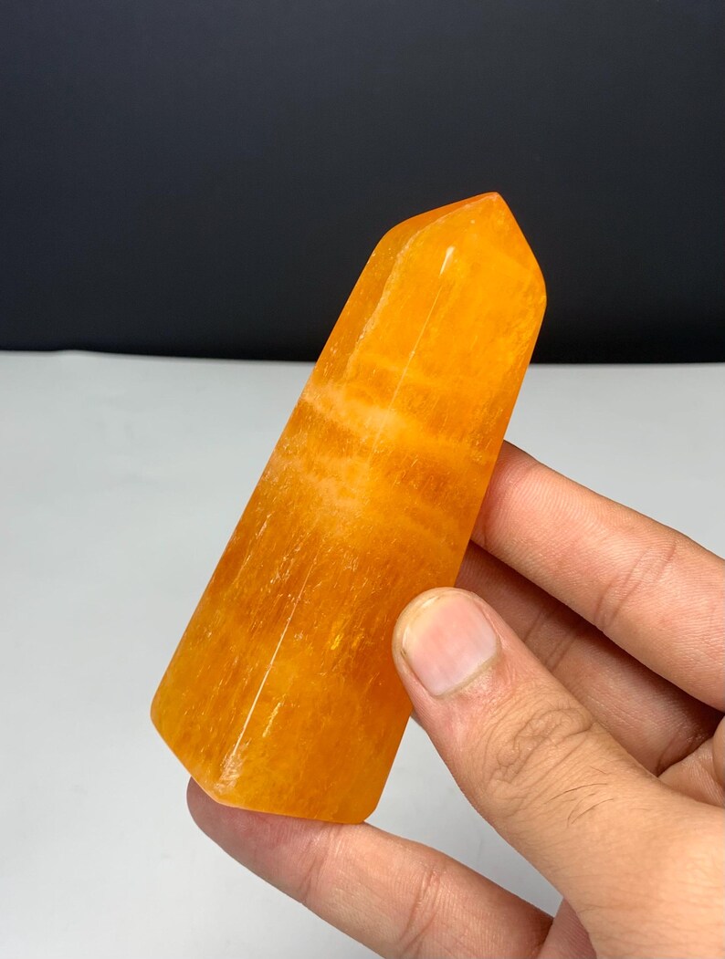 May include: A polished orange crystal tower with a hexagonal base and a pointed top. The stone features a translucent, vibrant orange hue with visible internal banding and natural mineral inclusions throughout its smooth, faceted surface.