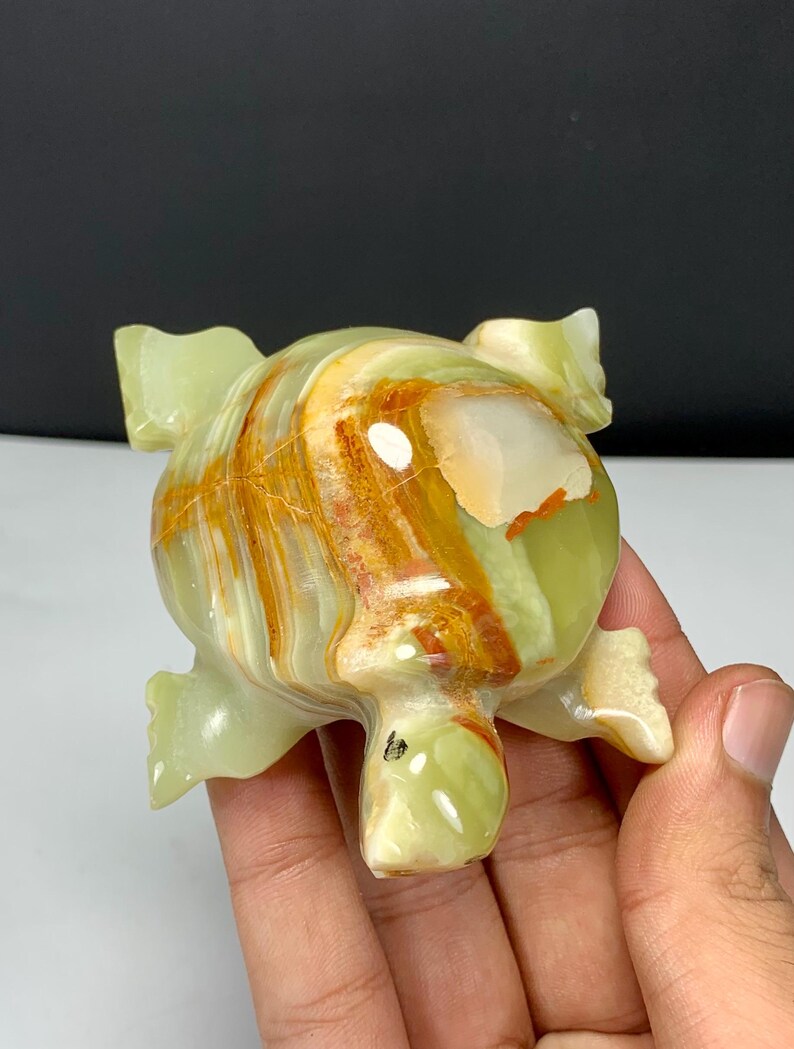 May include: A polished stone turtle figurine carved from light green onyx with prominent bands of orange and white mineral inclusions. The smooth, rounded shell features a glossy finish with visible natural veining and a distinct color gradient.