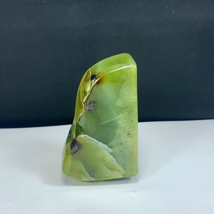 Pode incluir: Uma pedra polida de formato irregular em tons de verde claro e lima com áreas translúcidas. A superfície apresenta veios brancos naturais e pequenas inclusões castanho-escuras, exibindo uma textura lisa e brilhante.