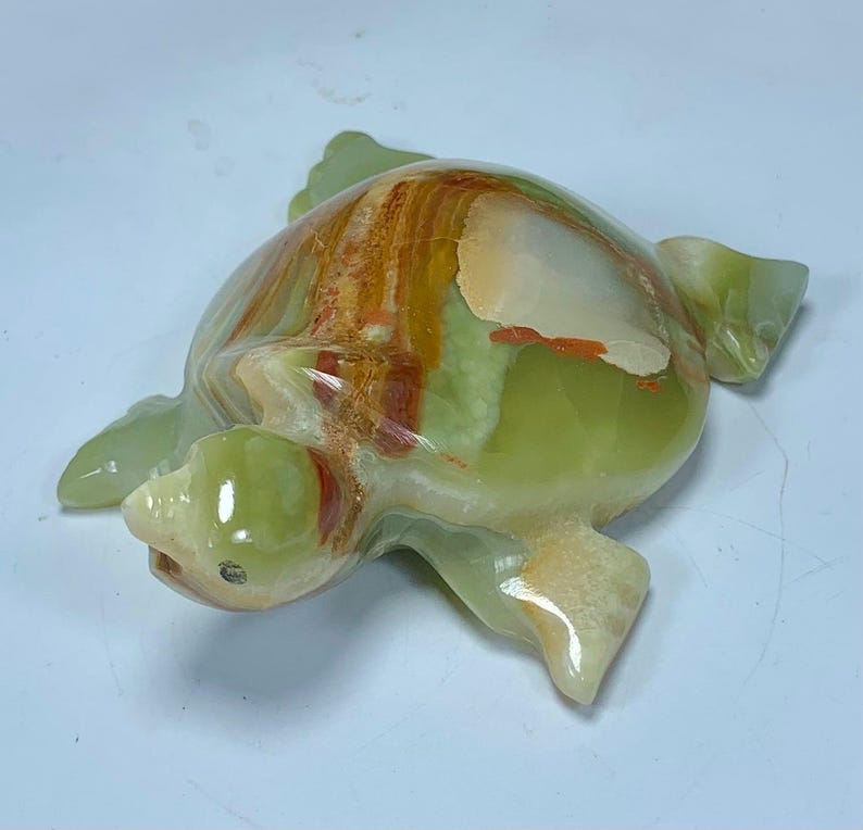 May include: A polished stone turtle figurine carved from translucent green onyx with natural banding in shades of cream, tan, and deep orange. The smooth, rounded shell features organic mineral patterns, while the head and flippers are sculpted with a glossy finish, showcasing the stone's unique color variations and light-reflecting properties.