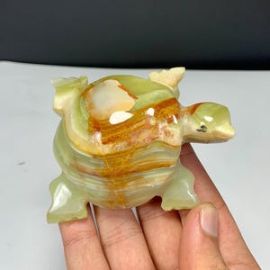 May include: A polished stone turtle figurine carved from banded green and orange onyx. The sculpture features a smooth, glossy surface with natural mineral layers, a rounded shell, four distinct legs, and a small head with a dark spot for an eye.