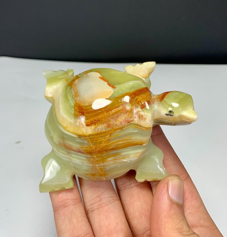 May include: A polished stone turtle figurine carved from banded green and orange onyx. The sculpture features a smooth, glossy surface with natural mineral layers, a rounded shell, four distinct legs, and a small head with a dark spot for an eye.