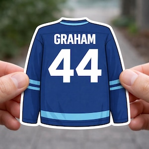 Könnte beinhalten: Ein blauer Eishockeytrikot-Aufkleber mit dem Namen GRAHAM in weißen Großbuchstaben über der Nummer 44. Das Trikot hat hellblaue horizontale Streifen an den Ärmeln und am unteren Saum sowie einen weißen Rand, der die gesamte Form umreißt.