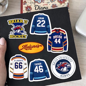May include: A collection of vinyl stickers featuring Briar U Hockey logos, a Malone's oval emblem, and four hockey jerseys with names and numbers including Logan 22, Briar 44, Di Laurentis 66, and Tucker 46, all displayed on a black spiral notebook.