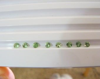 May include: Eight round, green gemstones arranged in a row on a white surface.