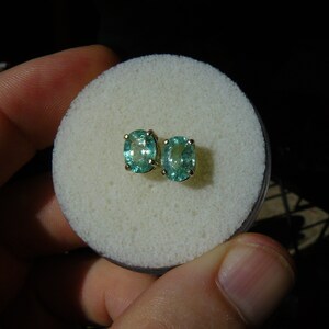 May include: A pair of oval-shaped green gemstone earrings with gold settings. The earrings are displayed on a white foam surface.