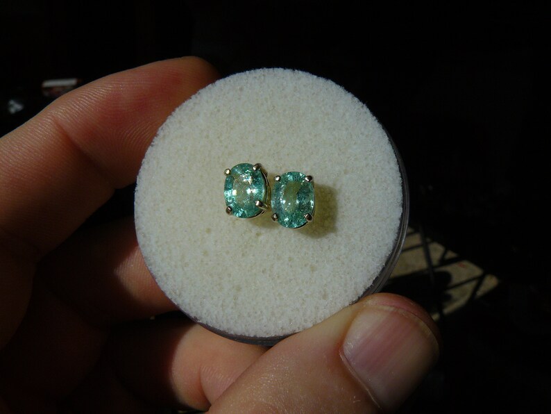 May include: A pair of oval-shaped green gemstone earrings with gold settings. The earrings are displayed on a white foam surface.