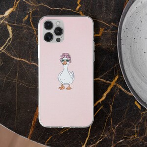 Pu&ograve; includere: Custodia per telefono rosa con un'oca dei cartoni animati che indossa occhiali da sole e un bigodino rosa. L'oca ha becco e zampe arancioni. La custodia ha un bordo trasparente e una fotocamera multi-obiettivo.