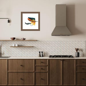 Pode incluir: Uma pintura em aquarela emoldurada de uma isca de pesca. A isca apresenta um anzol preto, com penas laranja, amarelas e azuis. A obra de arte é exibida em uma cozinha com armários de madeira e uma parede de azulejos brancos.