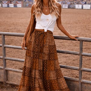 May include: A woman wearing a brown cowboy hat, white tank top, and a long, patterned brown skirt. She is also wearing cowboy boots. The background shows a rodeo arena with spectators.