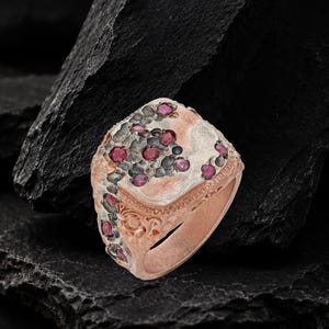 Puede incluir: Un anillo de metal en tono rosado con una superficie texturizada similar a un panal y acentos oscuros oxidados. El anillo está engastado con varias piedras preciosas pequeñas, redondas y de color rojo oscuro incrustadas en el diseño.