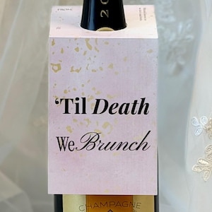 Könnte beinhalten: Ein rechteckiger Flaschenanhänger aus Papier in zartem Rosa mit goldenen Sprenkeln, der den schwarzen Text 'Til Death We Brunch' und 'FROM' oben trägt. Der Anhänger ist um den Hals einer dunklen Champagnerflasche mit der Jahreszahl 2015 platziert.