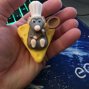 May include: A small, three-dimensional figurine of a grey mouse wearing a white chef hat, sitting on a yellow wedge of Swiss cheese. The mouse holds a brown wooden-style spoon in its front paws, featuring large black and white eyes and a white nose.