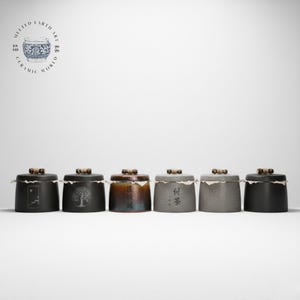 Puede incluir: Seis botes de té de cerámica con tapas oscuras y asas de madera. Los botes son de varios colores, incluyendo negro, gris y marrón, con diseños decorativos y caracteres chinos. Las tapas están selladas con papel.
