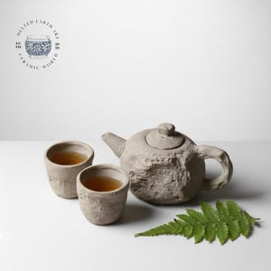 May include: A handcrafted ceramic tea set, featuring a textured teapot and two matching cups, filled with tea. The set is a neutral beige colour, with a fern leaf in the foreground. The logo "Melted Earth Art Ceramic World" is in the upper left corner.
