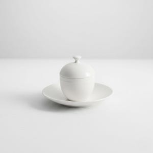 Peut inclure: Un sucrier en céramique blanche avec un couvercle et une petite poignée, posé sur une soucoupe assortie. Le bol a une forme arrondie et l'ensemble est entièrement blanc. Le fond est une surface blanche unie.