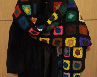 May include: A black coat with a colorful, crocheted scarf. The scarf features a repeating pattern of squares in various colors, including pink, yellow, green, and blue. The coat is long and appears to be made of a soft material.