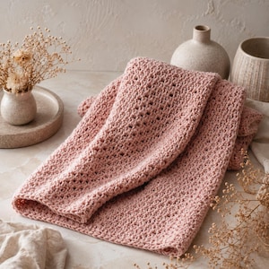 May include: A folded, dusty rose-colored crochet blanket. The blanket has a textured, open weave pattern. The image also includes a small vase with dried flowers, a decorative tray, and two ceramic vases in the background.