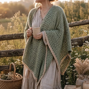 May include: A green crochet shawl with a light pink border is draped over a person. The shawl is worn over a light-colored dress. A basket of flowers and a mug are also visible. The background includes a wooden fence and wildflowers.
