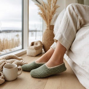 May include: A pair of sage green crochet slippers with a cream-colored trim. The slippers are on a wooden floor near a window. A cup of tea and a woven basket are also visible.